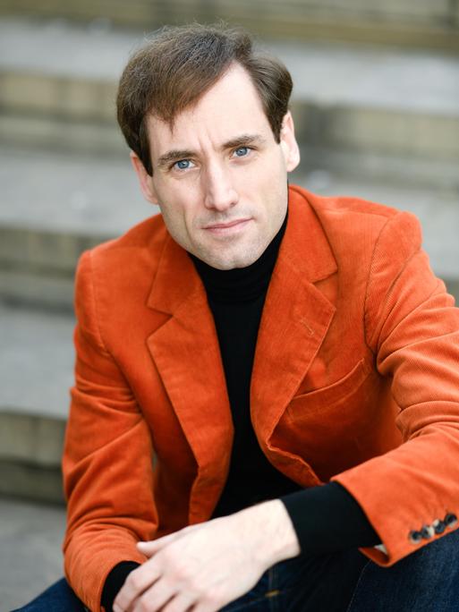 Ein Mann mit braunen Haaren und blauen Augen sitzt auf einer Treppe. Er schaut direkt in die Kamera. Er trägt ein orangefarbenes Cordjackett über einem schwarzen Shirt. 