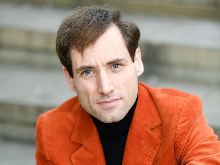 Ein Mann mit braunen Haaren und blauen Augen sitzt auf einer Treppe. Er schaut direkt in die Kamera. Er trägt ein orangefarbenes Cordjackett über einem schwarzen Shirt. 