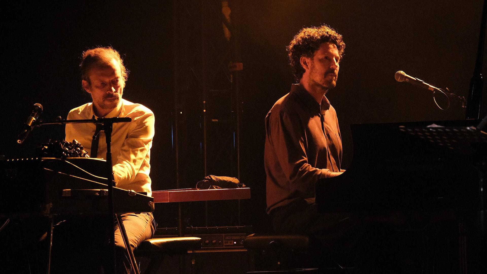 Zwei in sepiafarbenes Licht gehüllte Pianisten sitzen hinter ihren Instrumenten auf einer Bühne mit dunklem Hintergrund.