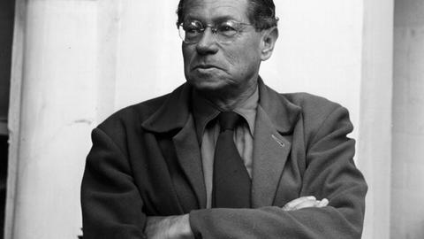 Portrait von Lion Feuchtwanger, der er eine Brille trägt und mit verschränkten Armen zur Seite schaut.