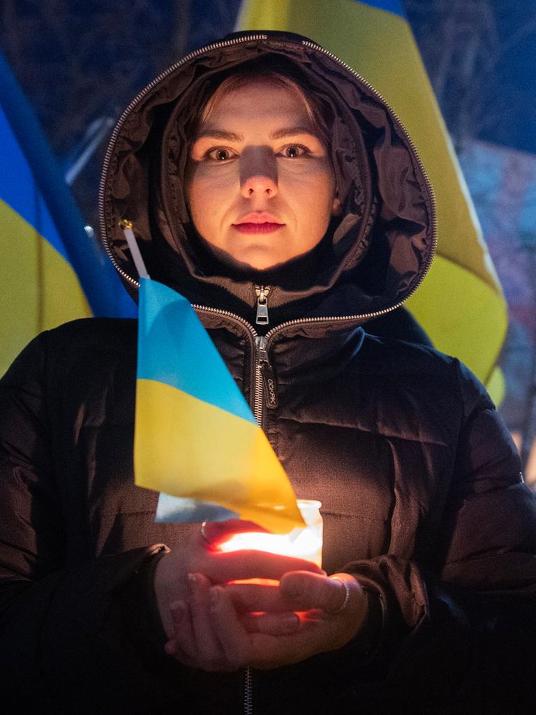 Zum Jahrestag des russischen Angriffs auf die Ukraine hält eine junge Frau bei einer Kundgebung in Montreal die ukranische Flagge und eine Kerze in der Hand, sie blickt in die Kamera. Im Hintergrund sind große ukrainische, gelb-blaue Flaggen zu sehen.