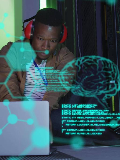 Symbolbild für Künstliche Intelligenz. Ein Mann mit Kopfhörer beugt sich in einem mit Computern vollgestellten Raum über eine dreidimensionale Grafik, die in grünen Umrissen ein menschliches Gehirn darstellt. 