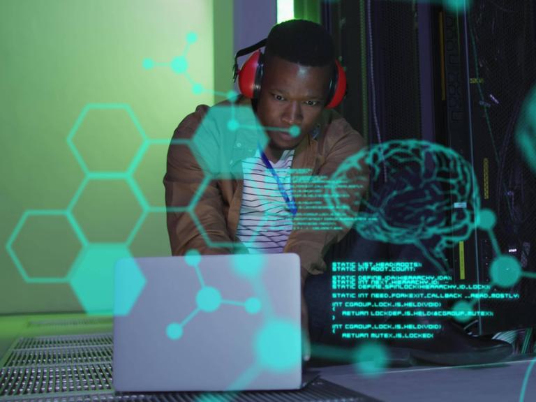 Symbolbild für Künstliche Intelligenz. Ein Mann mit Kopfhörer beugt sich in einem mit Computern vollgestellten Raum über eine dreidimensionale Grafik, die in grünen Umrissen ein menschliches Gehirn darstellt. 