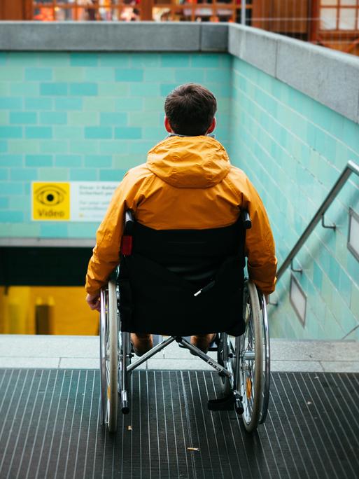 Ein Mann in einem Rollstuhl steht vor einer Treppe zu einem U-Bahnhofeingang.