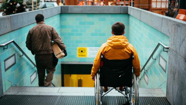 Ein Mann in einem Rollstuhl steht vor einer Treppe zu einem U-Bahnhofeingang.