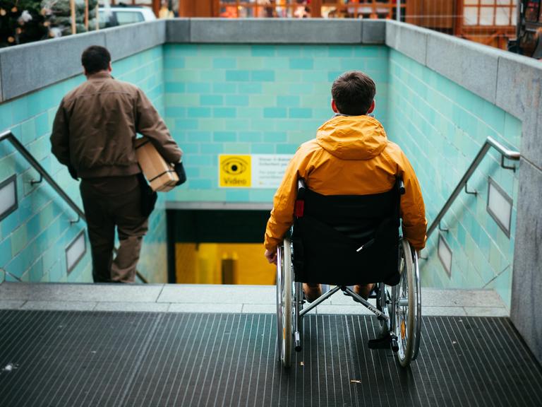 Ein Mann in einem Rollstuhl steht vor einer Treppe zu einem U-Bahnhofeingang.