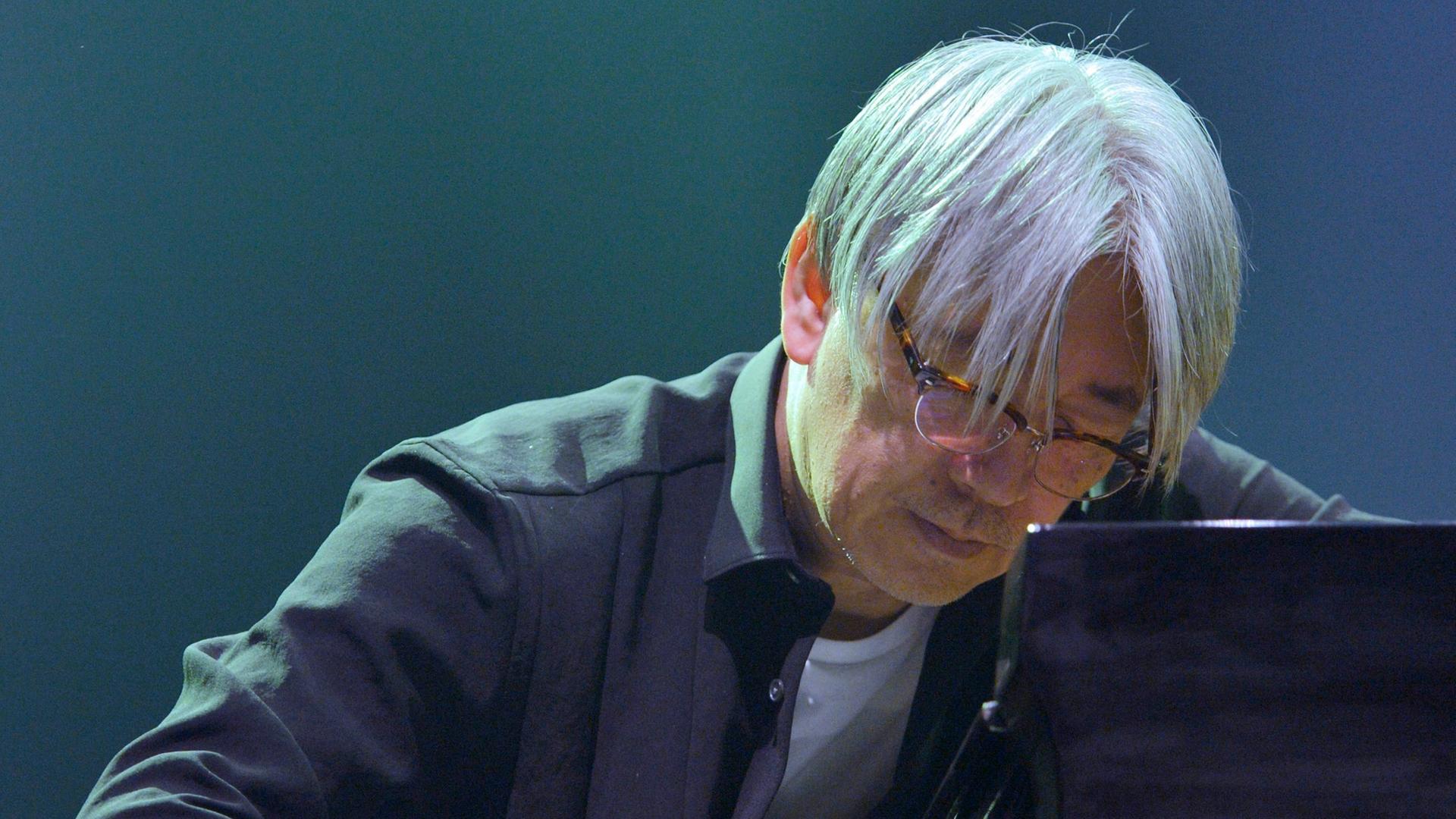 Der Pianist und Komponiest Ryuichi Sakamoto spielt auf einem Konzert Piano. Er hat den Kopf mit grauen Haaren weit an das Instrument hervorbegeugt. 