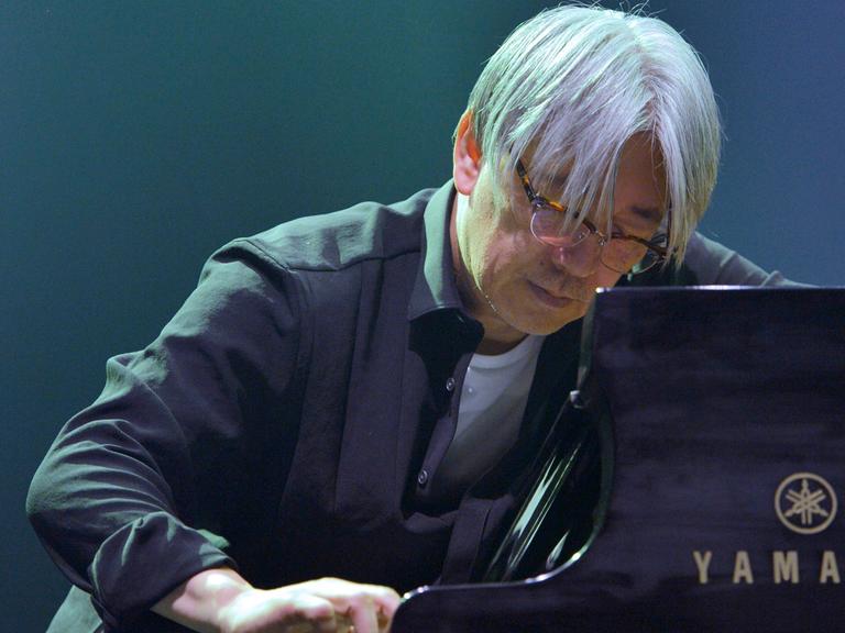 Der Pianist und Komponiest Ryuichi Sakamoto spielt auf einem Konzert Piano. Er hat den Kopf mit grauen Haaren weit an das Instrument hervorbegeugt. 