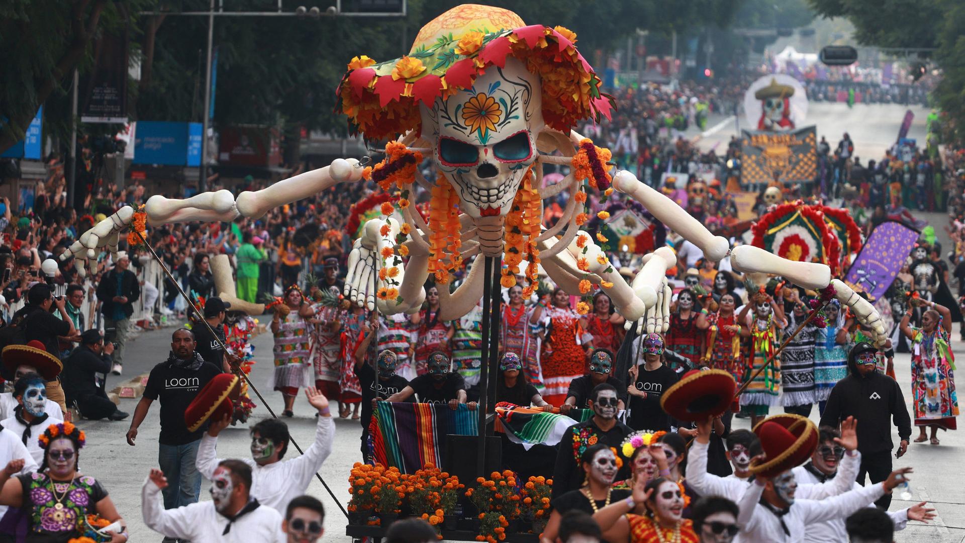 Eine Parade zum Tag der Toten in Mexiko. Menschen mit skelettähnlich-angemalten Gesichtern ziehen über eine Straße. Ein Wagen mit einem großen, bunt angemalten Skelett fährt in der Mitte. Das Skelett hat einen bunten Hut auf und die Arme ausgebereitet.