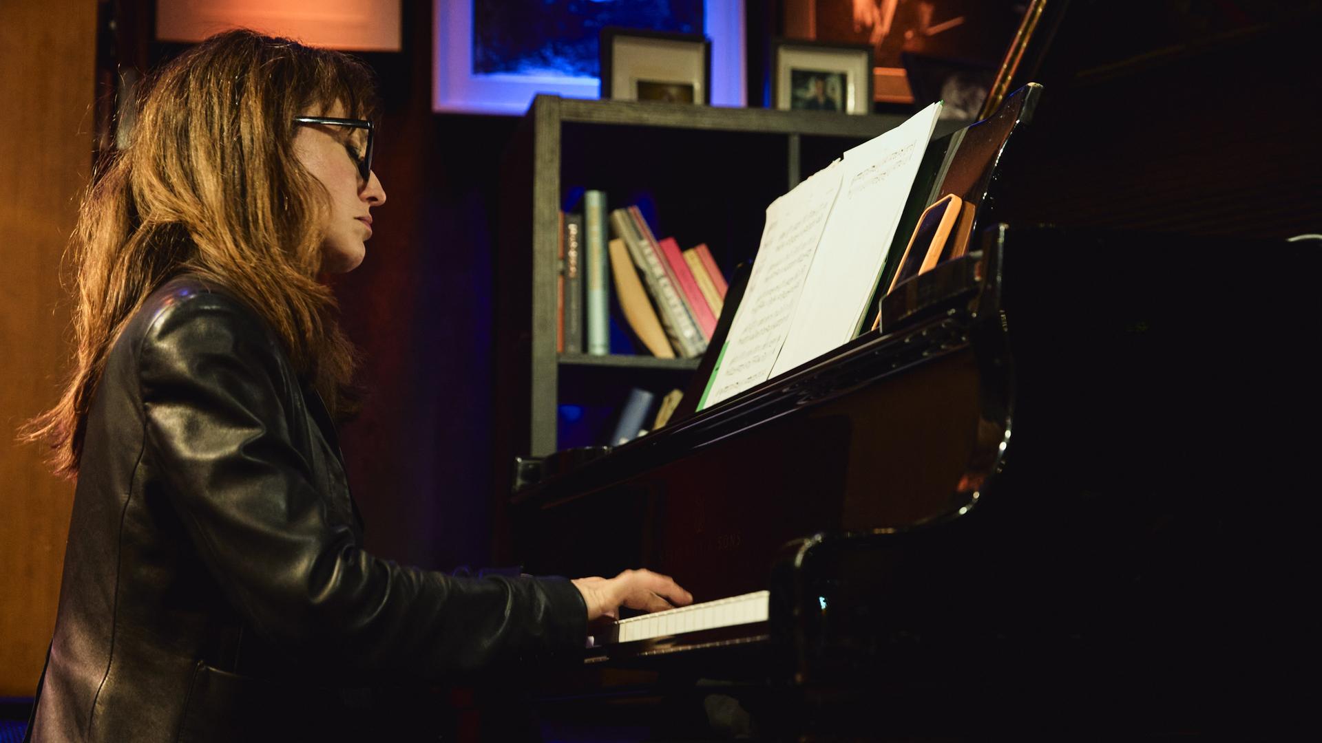 Marta Sánchez sitzt in einer Lederjacke mit offenem Haar und Brille in einer Clubumgebung an einem Flügel und spielt.