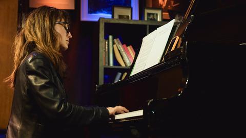 Marta Sánchez sitzt in einer Lederjacke mit offenem Haar und Brille in einer Clubumgebung an einem Flügel und spielt.