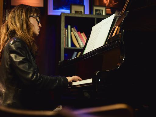 Marta Sánchez sitzt in einer Lederjacke mit offenem Haar und Brille in einer Clubumgebung an einem Flügel und spielt.