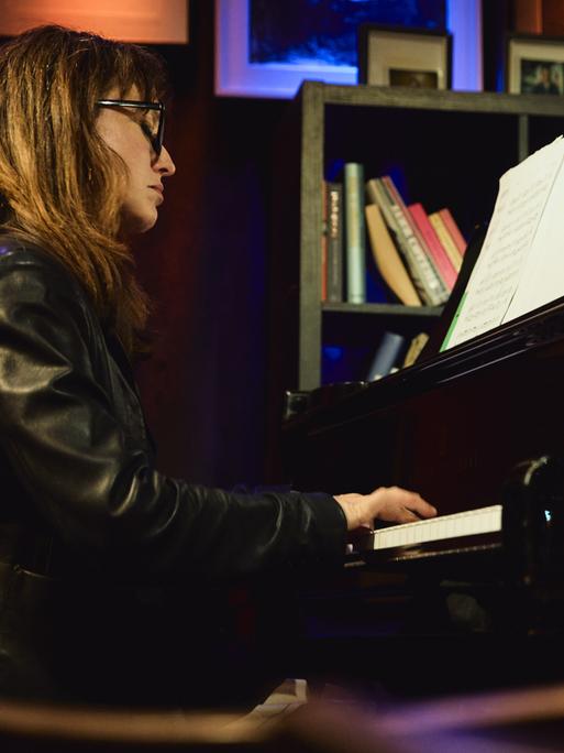 Marta Sánchez sitzt in einer Lederjacke mit offenem Haar und Brille in einer Clubumgebung an einem Flügel und spielt.