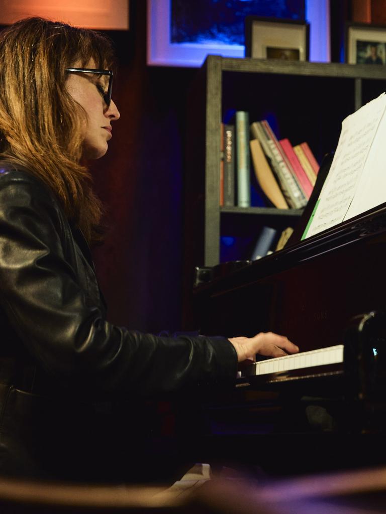 Marta Sánchez sitzt in einer Lederjacke mit offenem Haar und Brille in einer Clubumgebung an einem Flügel und spielt.