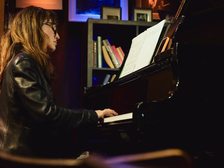 Marta Sánchez sitzt in einer Lederjacke mit offenem Haar und Brille in einer Clubumgebung an einem Flügel und spielt.