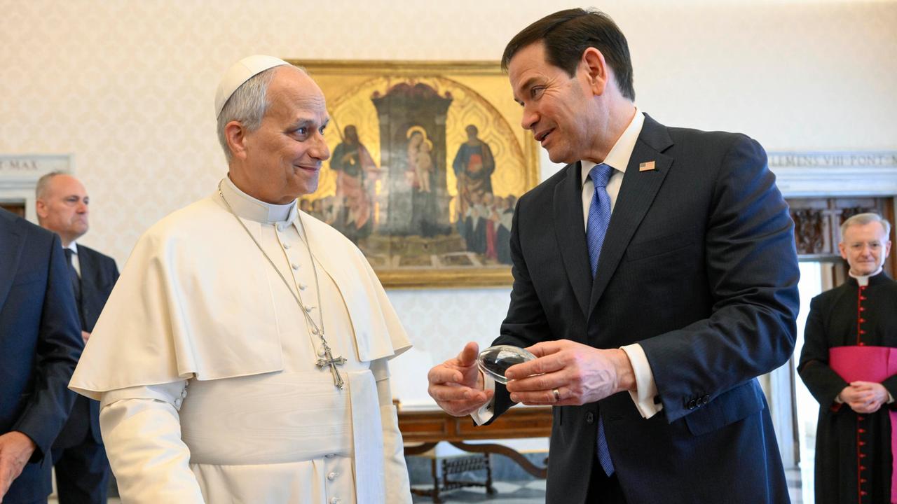 Die beiden stehen in einem Raum vor einer Vitrine und reden lächelnd miteinander. Rubio hält ein Exponat in der Hand. Er trägt ein schwarzes Jackett, der Papst im weißen Ornat.  Dahinter weitere Personen vor einer Wand mit einem Bild. 
