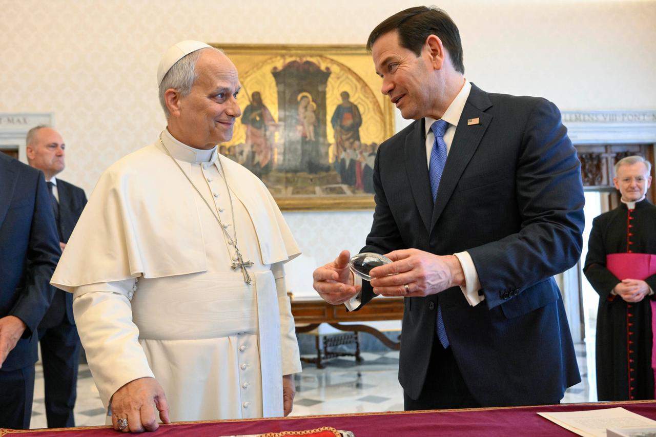 Die beiden stehen in einem Raum vor einer Vitrine und reden lächelnd miteinander. Rubio hält ein Exponat in der Hand. Er trägt ein schwarzes Jackett, der Papst im weißen Ornat.  Dahinter weitere Personen vor einer Wand mit einem Bild. 