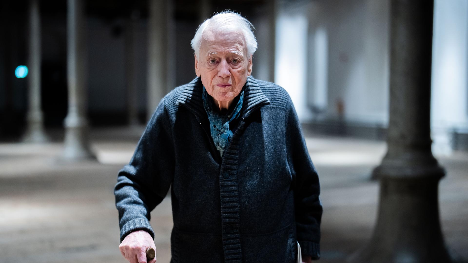 Alexander Kluge steht in einer Halle, er stützt sich auf seinem Stock und schaut direkt in die Kamera. Alexander Kluge steht in einer Halle, er stützt sich auf seinem Stock und schaut direkt in die Kamera.