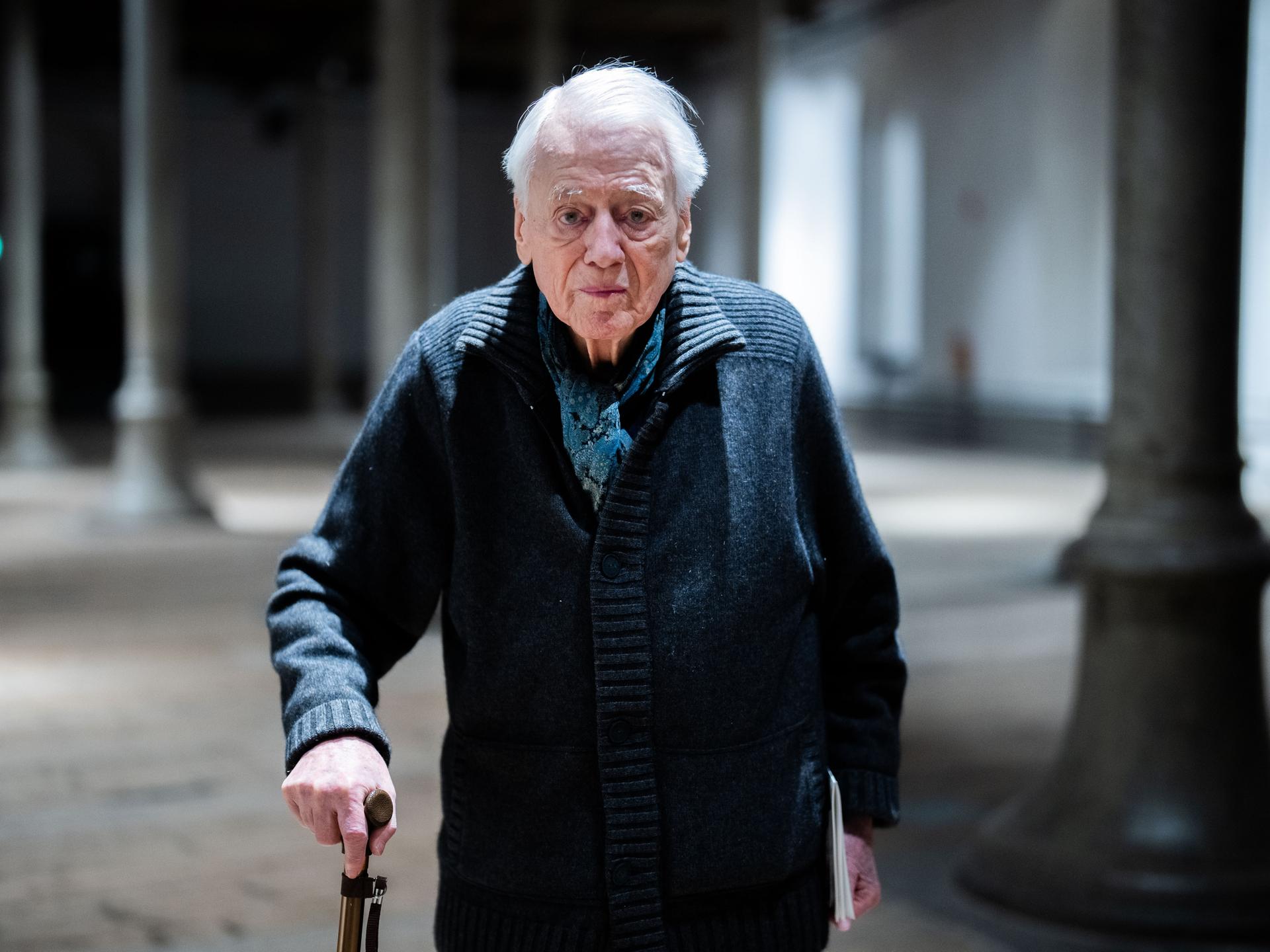 Alexander Kluge steht in einer Halle, er stützt sich auf seinem Stock und schaut direkt in die Kamera.