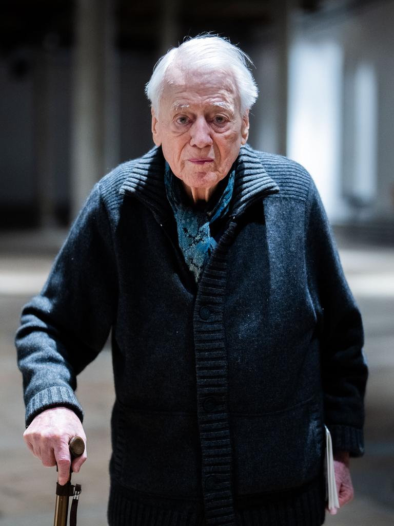 Alexander Kluge steht in einer Halle, er stützt sich auf seinem Stock und schaut direkt in die Kamera.