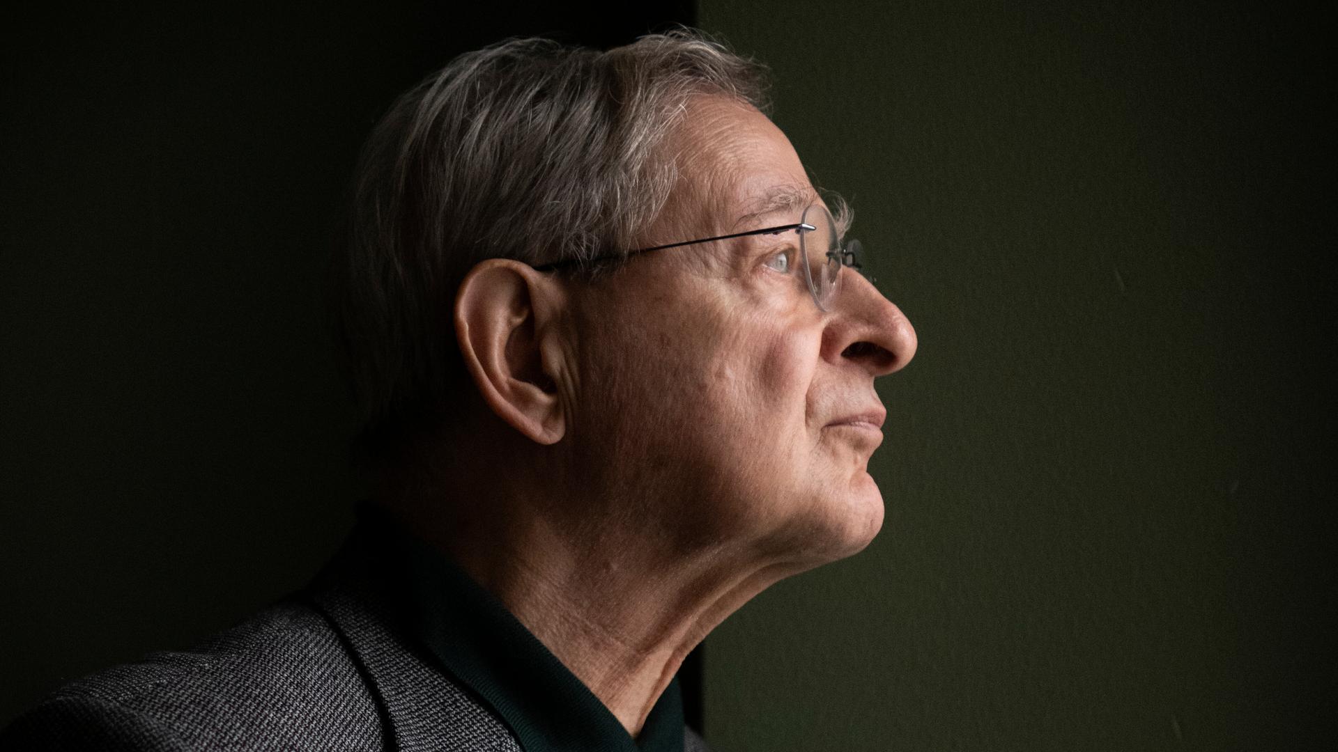 Péter Nádas steht im Profil vor einer dunklen Wand. Er trägt eine Brille und ein graues Sakko.