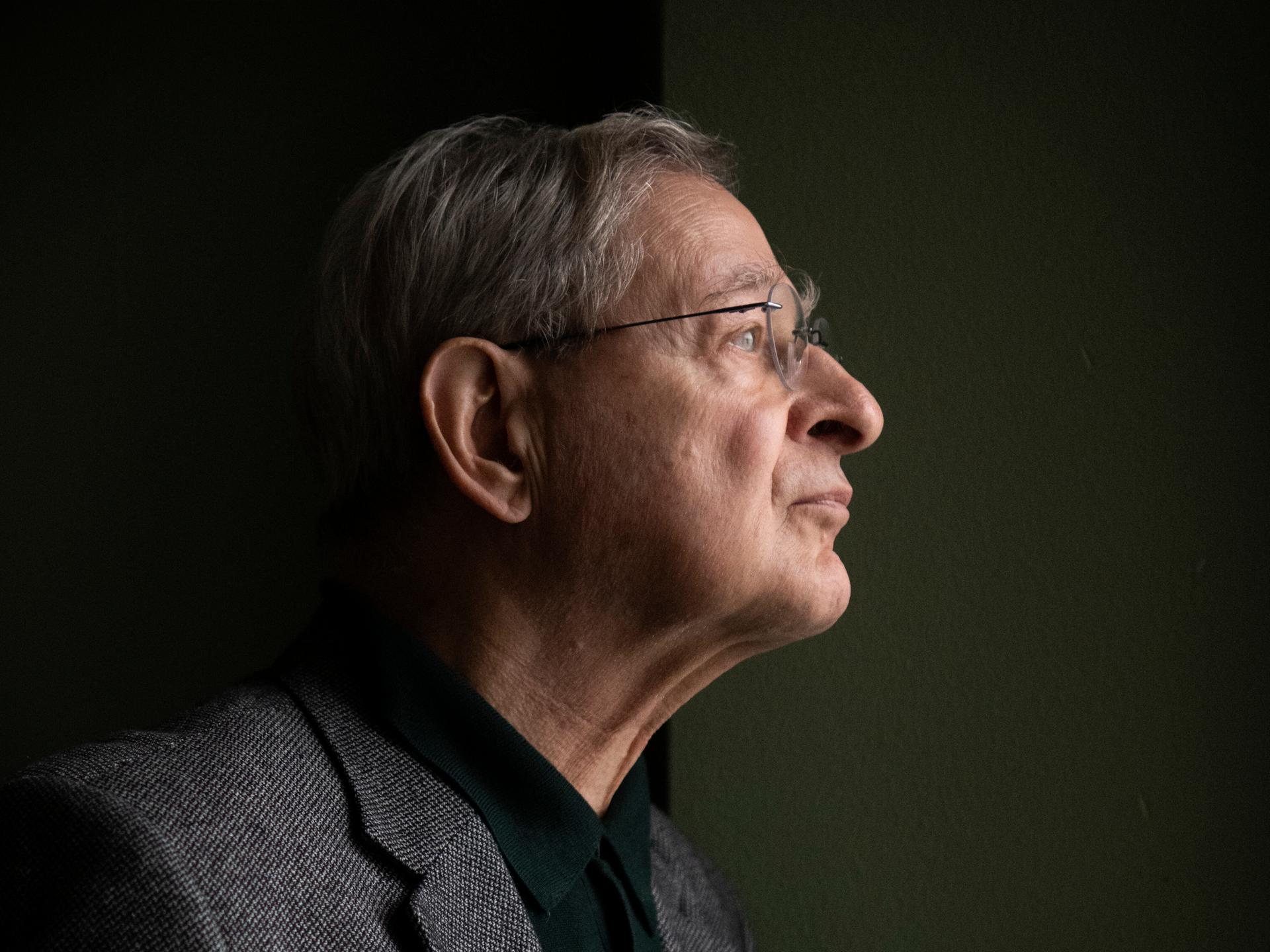 Péter Nádas steht im Profil vor einer dunklen Wand. Er trägt eine Brille und ein graues Sakko. Péter Nádas steht im Profil vor einer dunklen Wand. Er trägt eine Brille und ein graues Sakko.