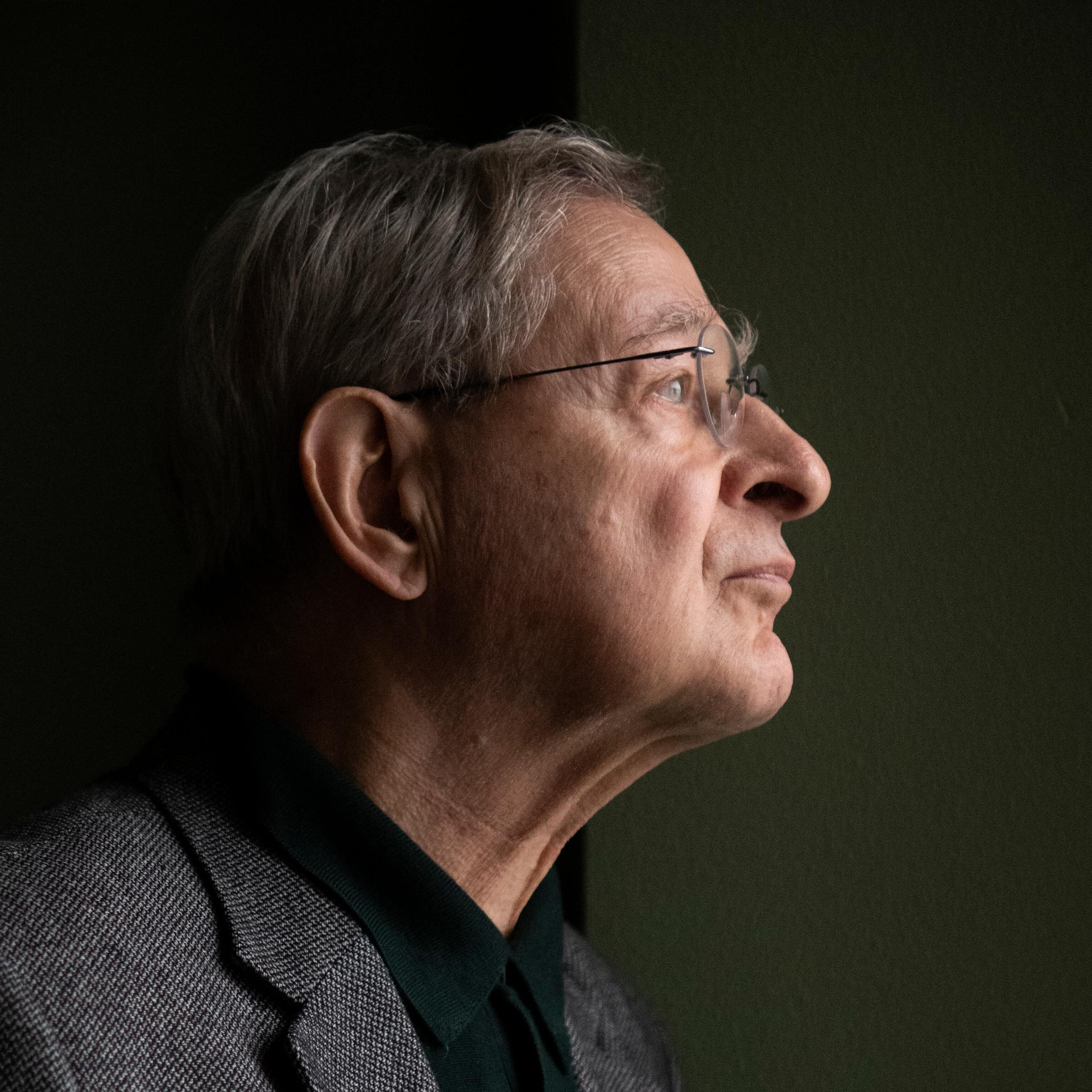 Péter Nádas steht im Profil vor einer dunklen Wand. Er trägt eine Brille und ein graues Sakko.