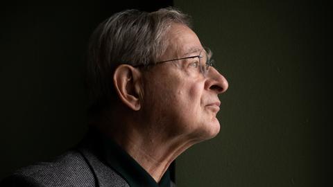 Péter Nádas steht im Profil vor einer dunklen Wand. Er trägt eine Brille und ein graues Sakko. Péter Nádas steht im Profil vor einer dunklen Wand. Er trägt eine Brille und ein graues Sakko.