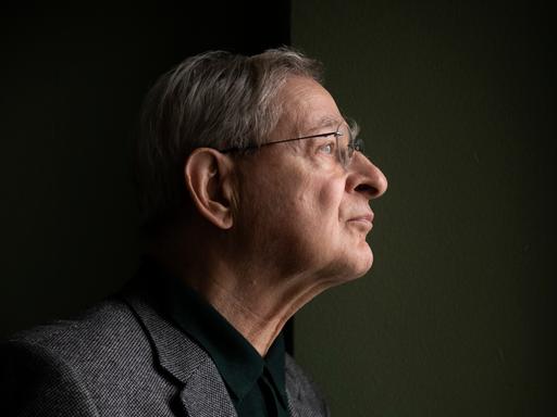 Péter Nádas steht im Profil vor einer dunklen Wand. Er trägt eine Brille und ein graues Sakko.