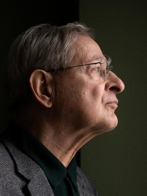 Péter Nádas steht im Profil vor einer dunklen Wand. Er trägt eine Brille und ein graues Sakko. Péter Nádas steht im Profil vor einer dunklen Wand. Er trägt eine Brille und ein graues Sakko.