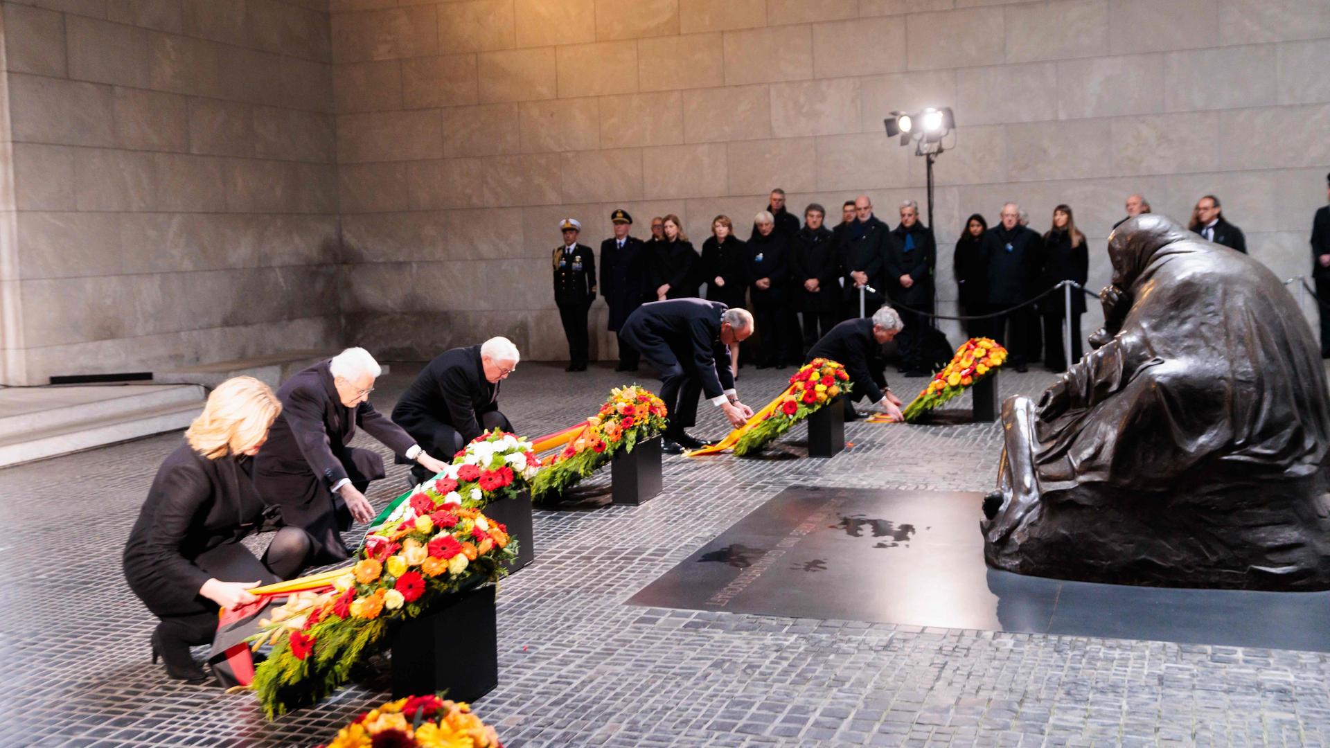 Die vier knien gleichzeitig vor runden Kränzen und zupfen an den Bändern. Dahinter stehend weitere Teilnehmer. Am rechten Bildrand eine große Metallskulptur. Die vier knien gleichzeitig vor runden Kränzen und zupfen an den Bändern. Dahinter stehend weitere Teilnehmer. Am rechten Bildrand eine große Metallskulptur.