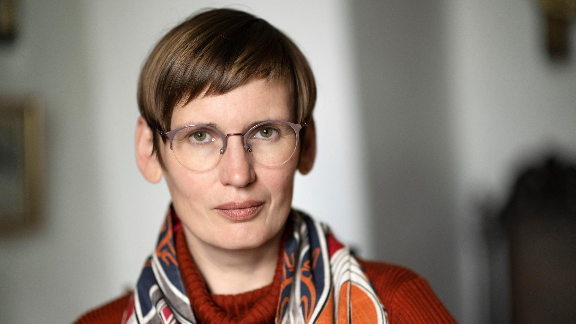 Eine Frau mit dunkelblonder Kurzhaarfrisur und runder Brille schaut nachdenklich in die Kamera. Sie trägt einen orangefarbenen Rollkragenpullover und einen bunt gemusterten Schal. Eine Frau mit dunkelblonder Kurzhaarfrisur und runder Brille schaut nachdenklich in die Kamera. Sie trägt einen orangefarbenen Rollkragenpullover und einen bunt gemusterten Schal.