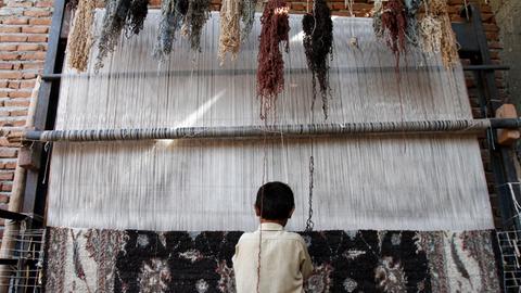 Ein Kind sitzt an einem Webstuhl und stellt einen Teppich her.