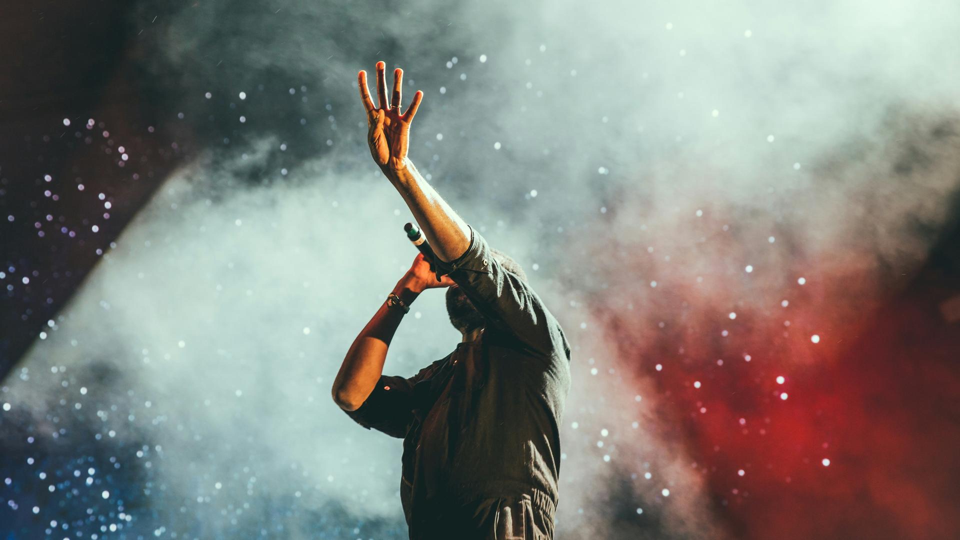Ein Mann in einem Pullover steht auf der Bühne mit Mikrophon, sein Gesicht ist durch seinen gehobenen Arm verdeckt, während der Bühnenhintergrund weiß und rot mit Nebel ausgeleuchtet ist.