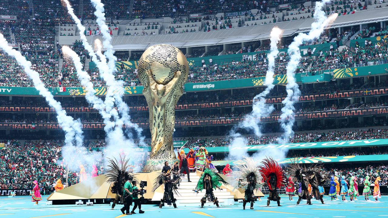 Einblicke in die Eröffnugsfeier der Fußball-WM. Menschen in Kostümen nehmen an der Show im Inneren des Stadions teil. Zu sehen ist ein großer WM-Pokal. 