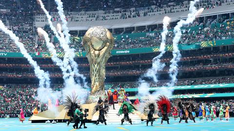 Einblicke in die Eröffnugsfeier der Fußball-WM. Menschen in Kostümen nehmen an der Show im Inneren des Stadions teil. Zu sehen ist ein großer WM-Pokal. 