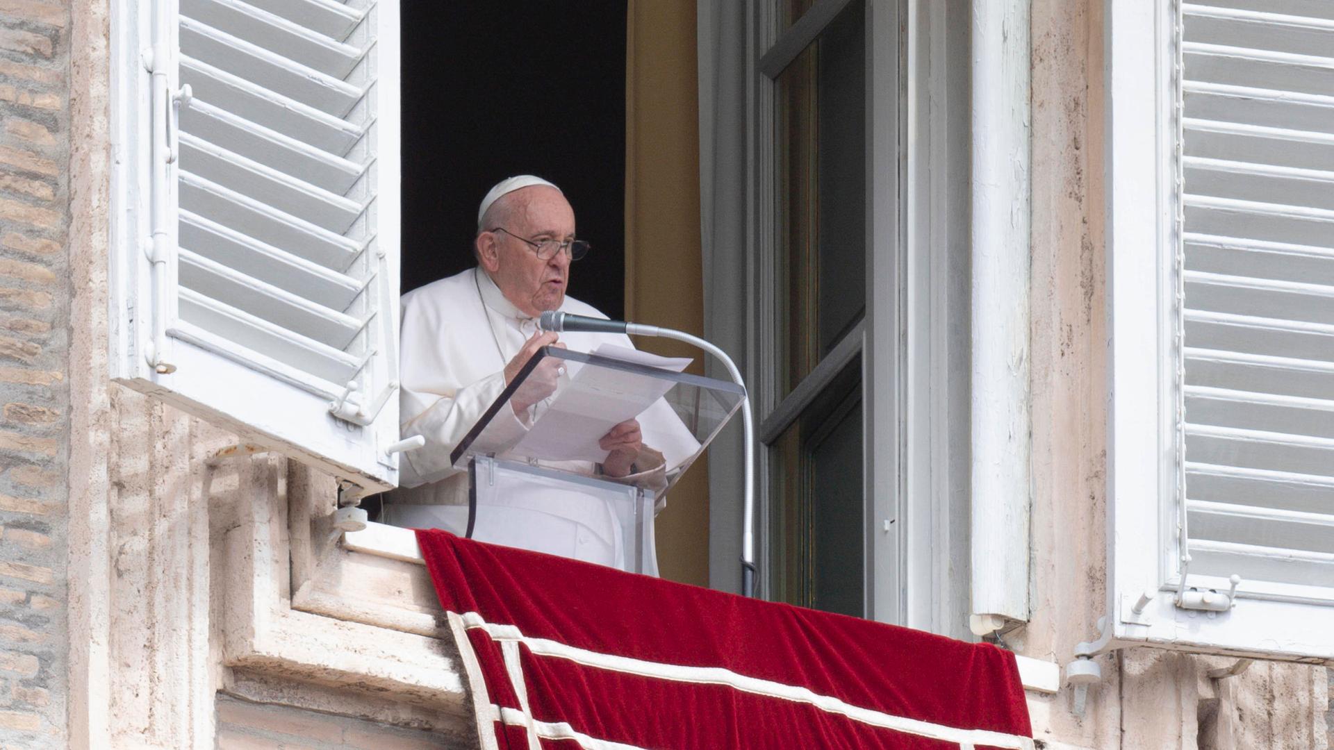 Papst Franziskus steht in weißem Gewand an einem durchsichtigen Rednerpult mit Mikrofon an einem offenen Fenster und spricht mit mehreren Seiten Papier in den Händen.