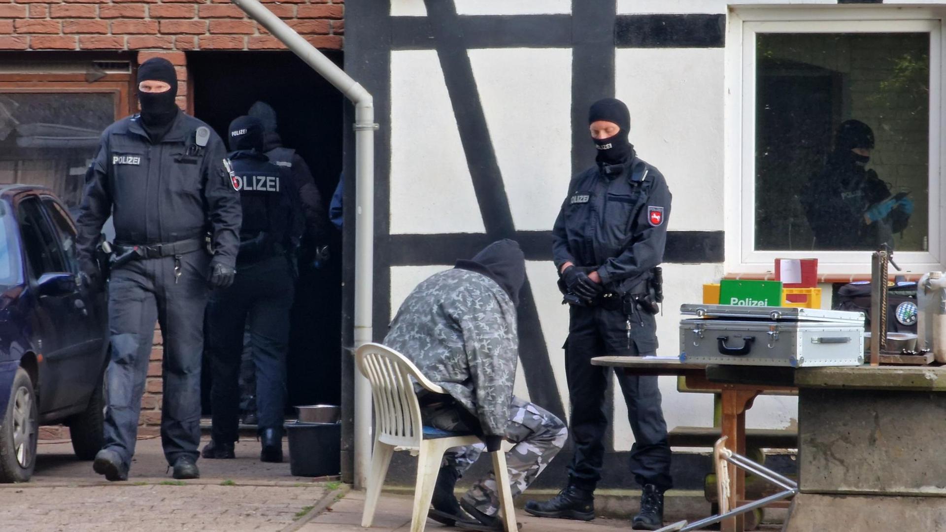 Nordrhein-Westfalen, Espelkamp: Polizisten sind bei einer Razzia im Einsatz, während ein Mann in Kleidung mit Tarnfleckmuster auf einem Stuhl vor einem Polizisten sitzt. D