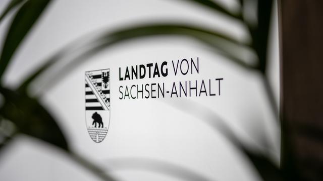 Das Logo des Landtags von Sachsen-Anhalt ist auf einem Schild zu sehen. Unscharfe Blätter im Vordergrund verdecken das Schild teilweise und bilden einen Rahmen um die Aufnahme. (Symbolbild)