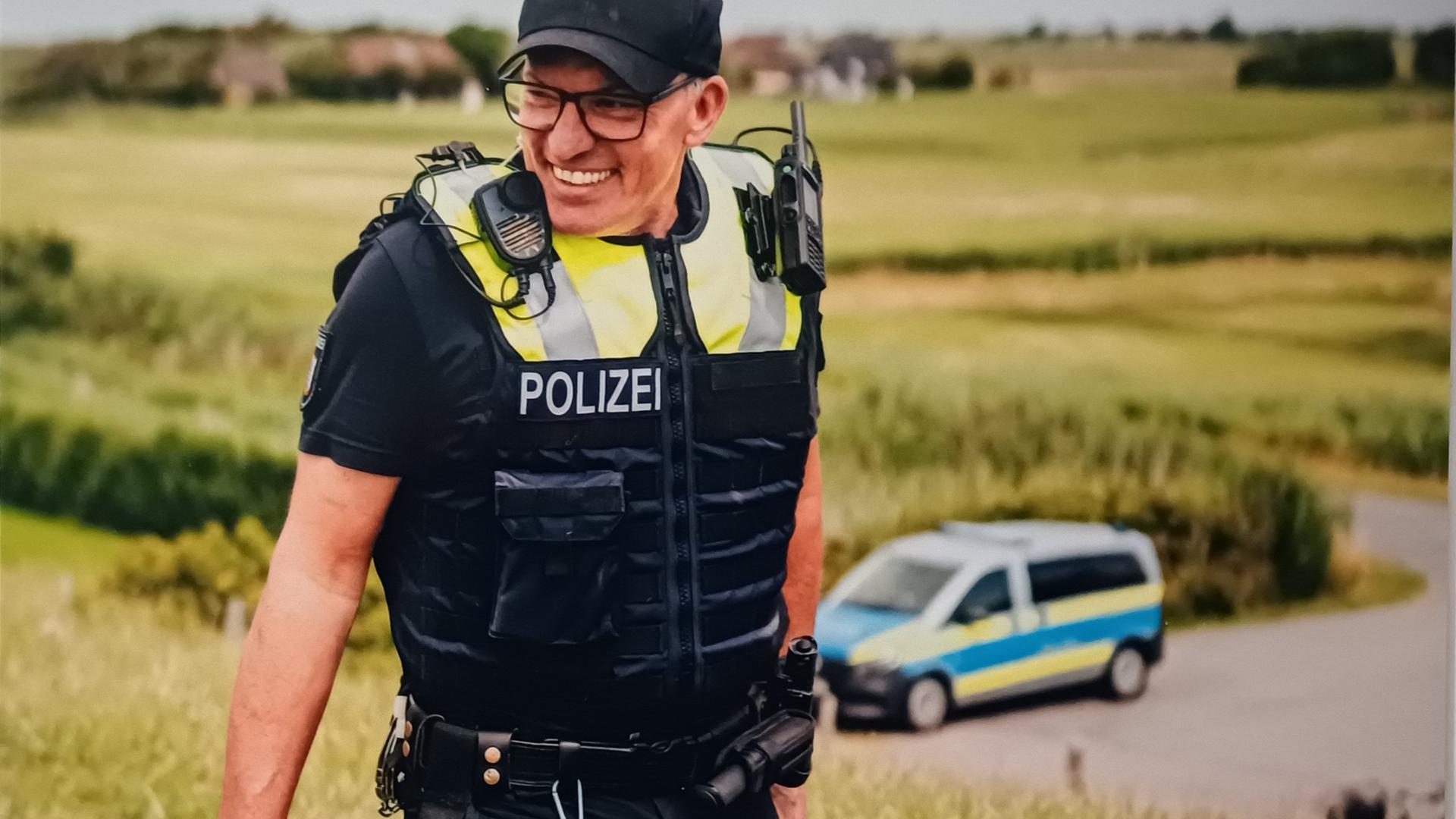 Der Inselpolizist Markus Stephan auf Pellworm, er ist in Uniform und lacht, im Hintergrund steht ein Polizeiauto. Der Inselpolizist Markus Stephan auf Pellworm, er ist in Uniform und lacht, im Hintergrund steht ein Polizeiauto.