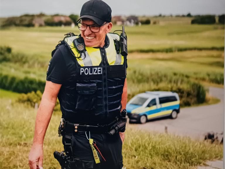 Der Inselpolizist Markus Stephan auf Pellworm, er ist in Uniform und lacht, im Hintergrund steht ein Polizeiauto.