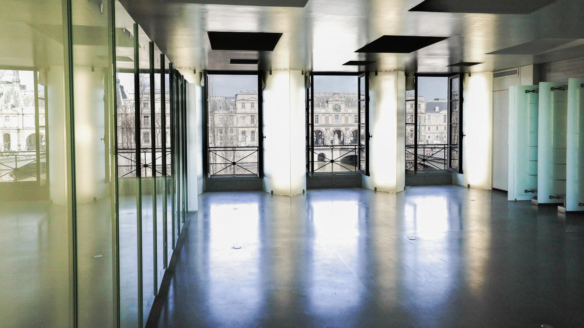 Auf dem Bild sieht man das Wohnzimmer von Karl Lagerfelds Pariser Wohnung. Ein heller, leerer Raum mit drei großen Fenstern, Steinboden und hellgrünen Glaselementen an der Seite. Durch die Fenster sieht man das Louvre-Museum.