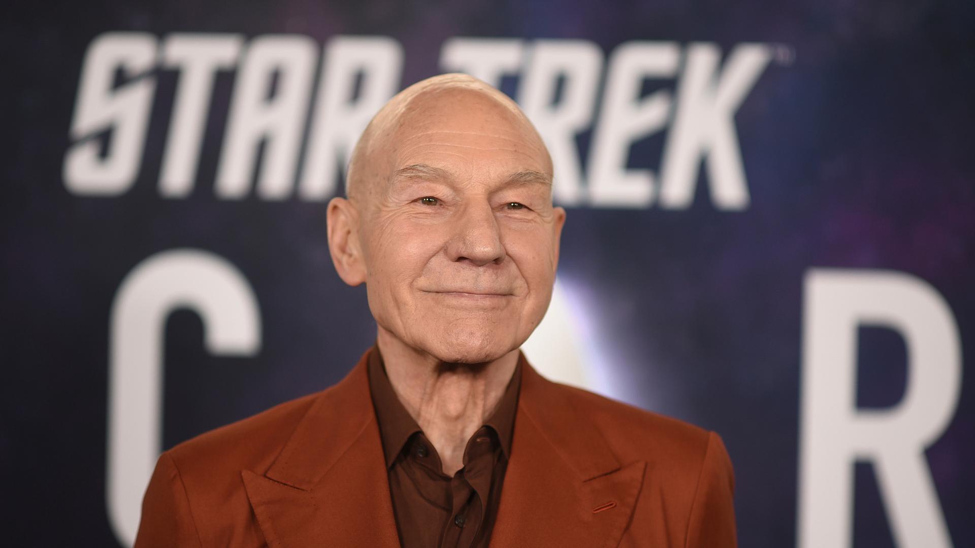 Stewart steht in einem braunen Sakko vor einem "Star Treck"-Plakat und lächelt. Man sieht nur seinen Oberkörper. 