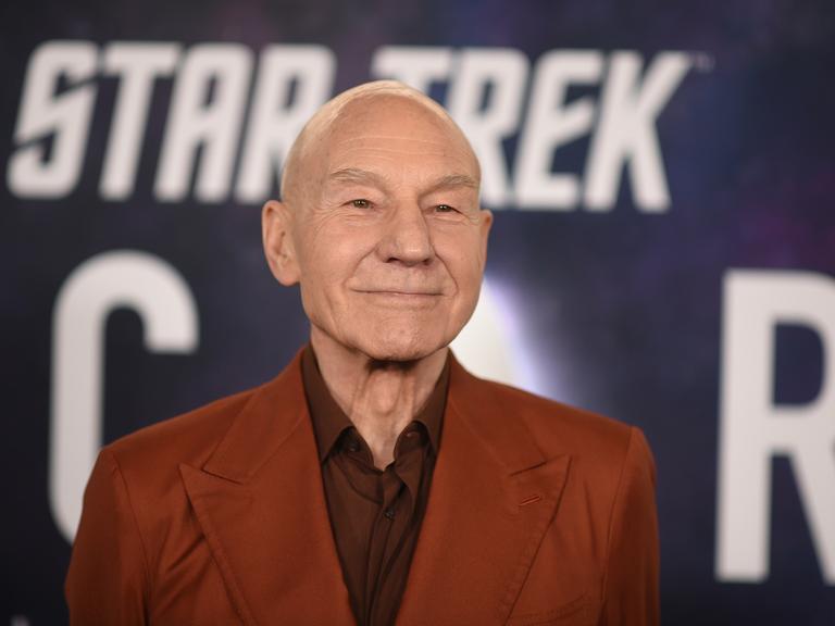 Stewart steht in einem braunen Sakko vor einem "Star Treck"-Plakat und lächelt. Man sieht nur seinen Oberkörper. 