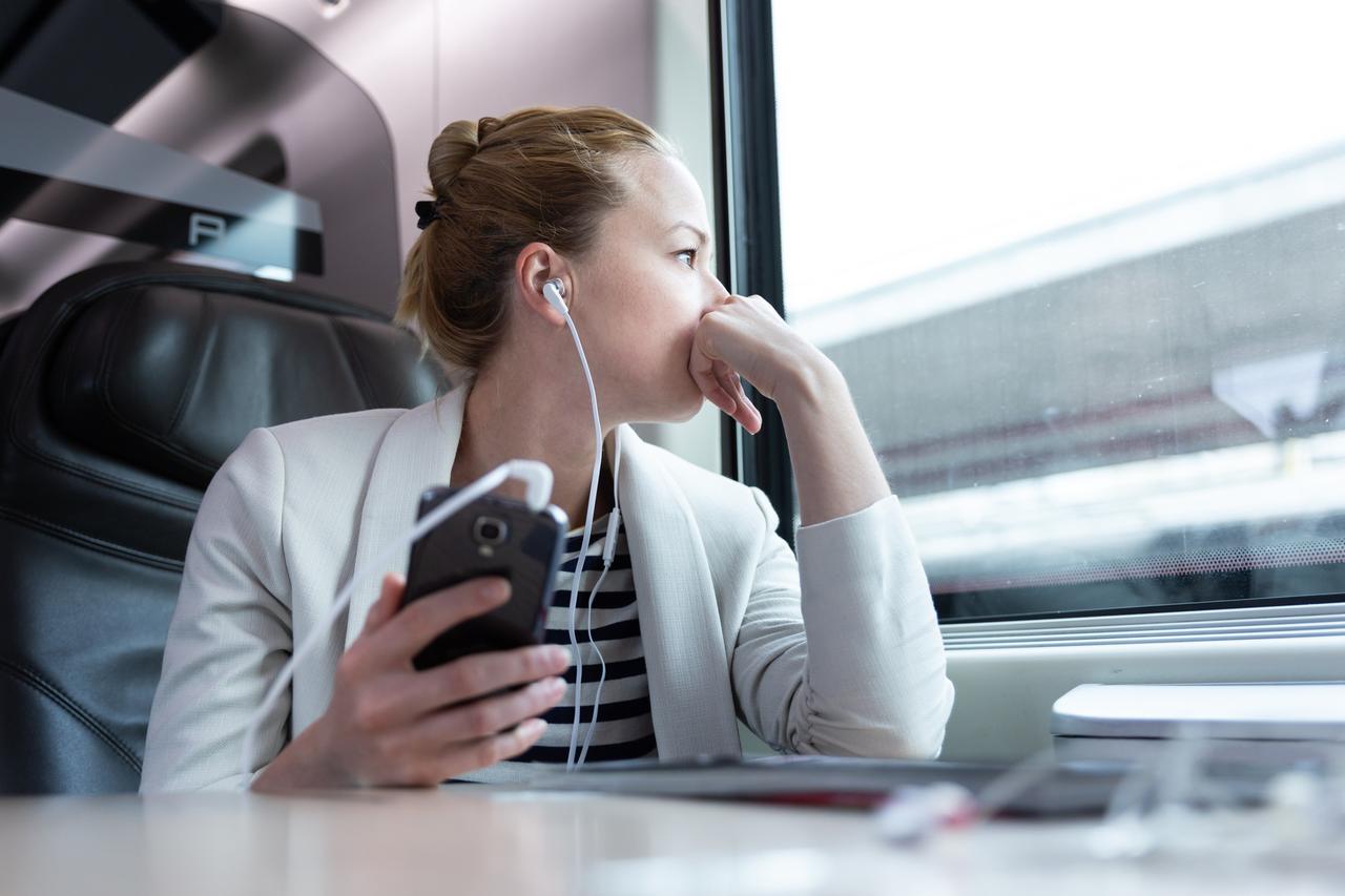 Eine Frau sitzt in der Bahn am Fenster mit Kopfhörern in den Ohren, die mit ihrem Handy verbunden sind.