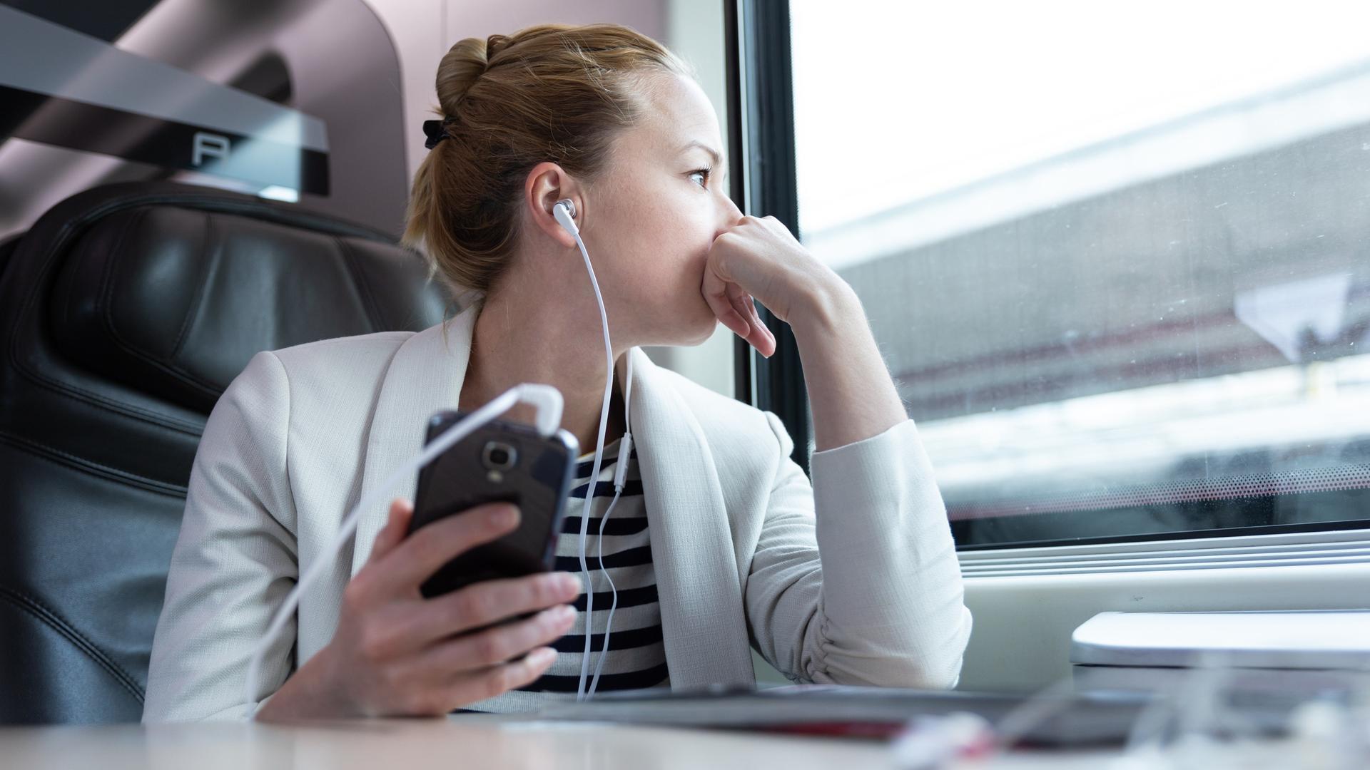 Eine Frau sitzt in der Bahn am Fenster mit Kopfhörern in den Ohren, die mit ihrem Handy verbunden sind.