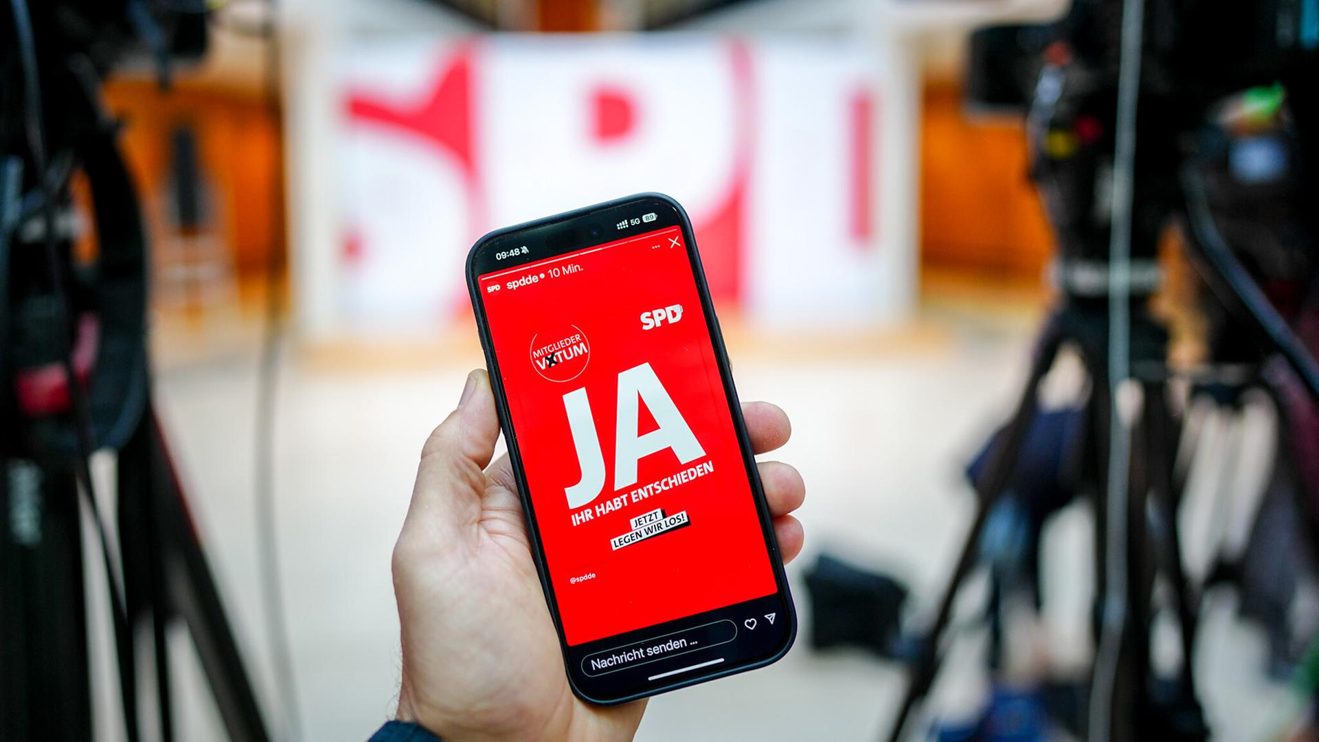 Auf einem Handy-Bildschirm ist auf einem roten Hintergrund das Wort „Ja“ zu lesen. Im Hintergrund sieht man in der Unschärfe einige aufgebaute Kameras für eine Pressekonferenz in der SPD-Parteizentrale. Auf einem Handy-Bildschirm ist auf einem roten Hintergrund das Wort „Ja“ zu lesen. Im Hintergrund sieht man in der Unschärfe einige aufgebaute Kameras für eine Pressekonferenz in der SPD-Parteizentrale.