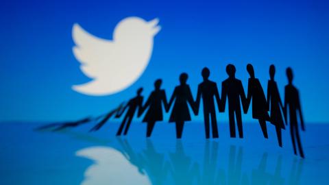 Das Twitter-Logo - der weiße Vogel - vor blauem Hintergrund, auf dem in der Ferne schwarze nebeneinander stehende Silhouetten erkennbar sind, die der Reihe nach umkippen.