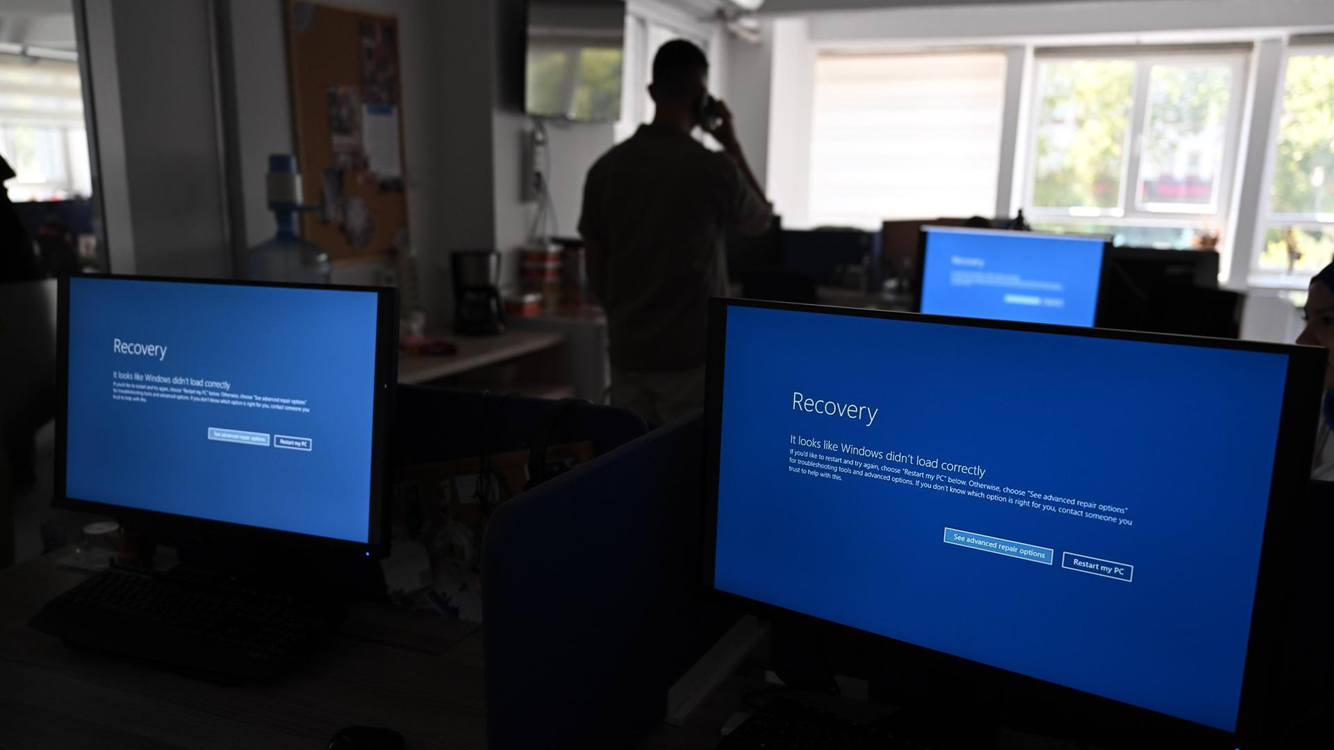 In einem Büro sind drei Monitore zu sehen, deren Display blau ist. Zudem ist zu erkennen, dass ein Mensch im dunkleren Teil des Raums steht.