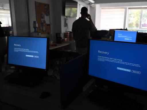 In einem Büro sind drei Monitore zu sehen, deren Display blau ist. Zudem ist zu erkennen, dass ein Mensch im dunkleren Teil des Raums steht.